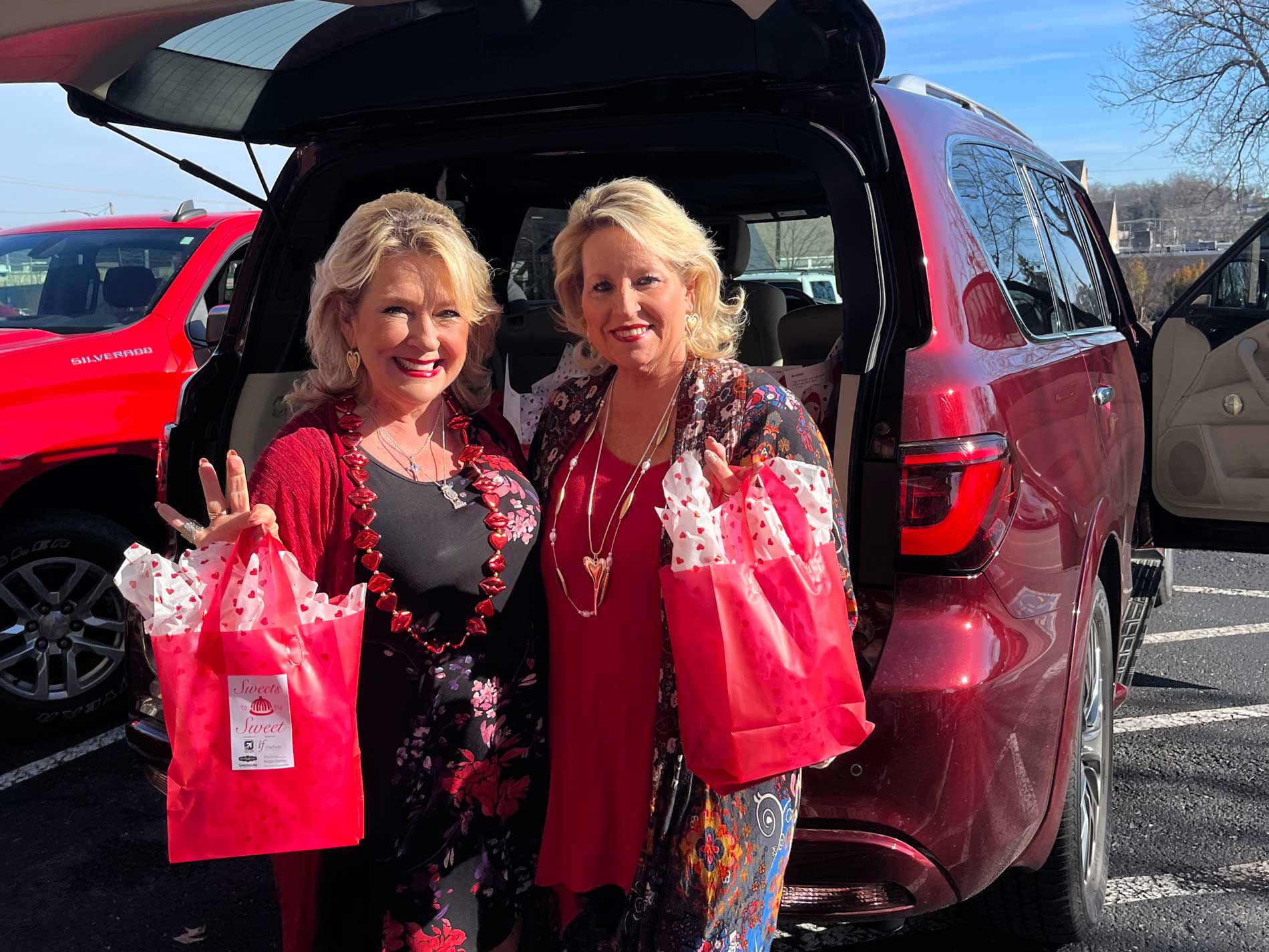 Two women holding pink bags in front of the trunk of a car.